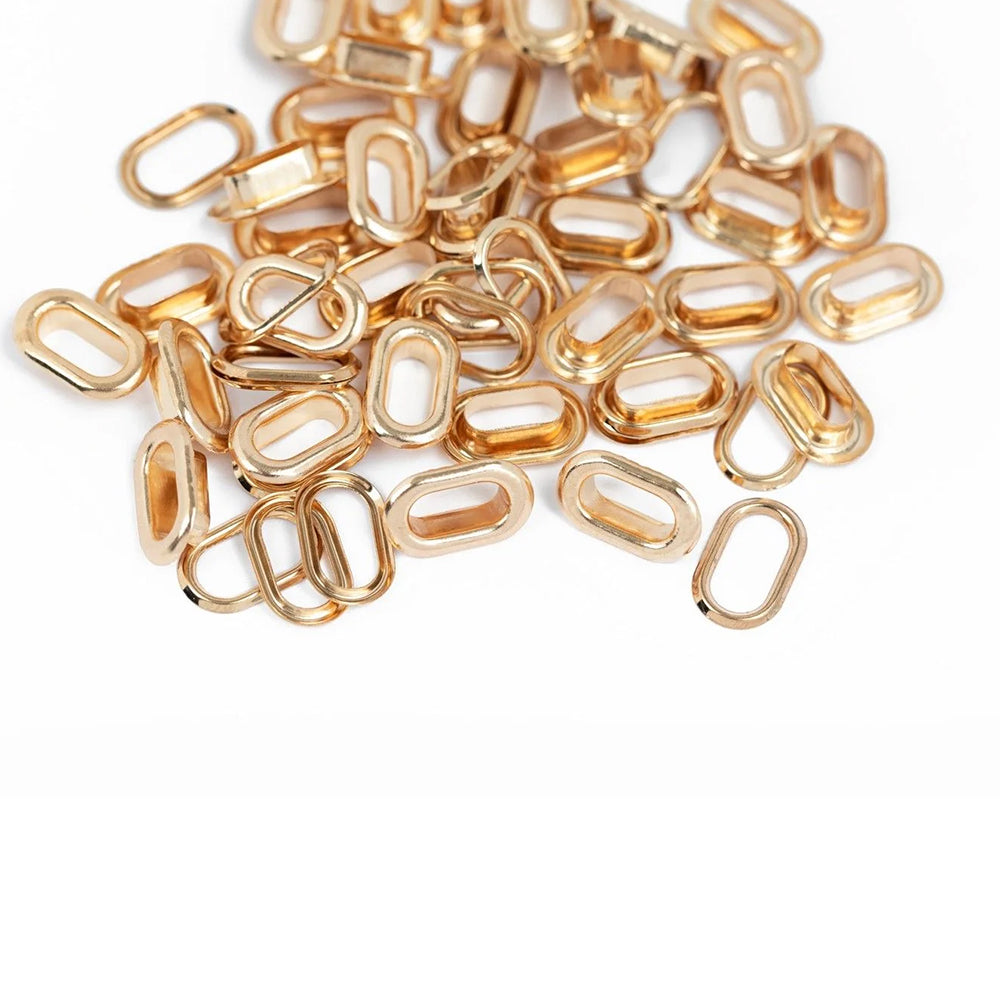 Ein Stapel kleiner, goldfarbener, ovaler Metallglieder von Vaessen liegt auf einer weißen Unterlage – ideal für Scrapbooking-Projekte. Diese Glieder, die perfekt mit den „Oval Crop-A-Dile“-Werkzeugen kompatibel sind und oft als „Metall-Ösen“ bezeichnet werden, verleihen jedem kreativen Projekt einen Hauch von Eleganz.