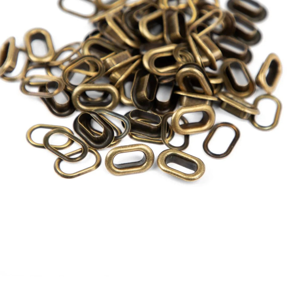 Auf einem weißen Hintergrund liegt ein Stapel kleiner, messingfarbener „Vaessen Metall-Eyelets“ – ovale Ringe, die sich perfekt für das Scrapbooking eignen.