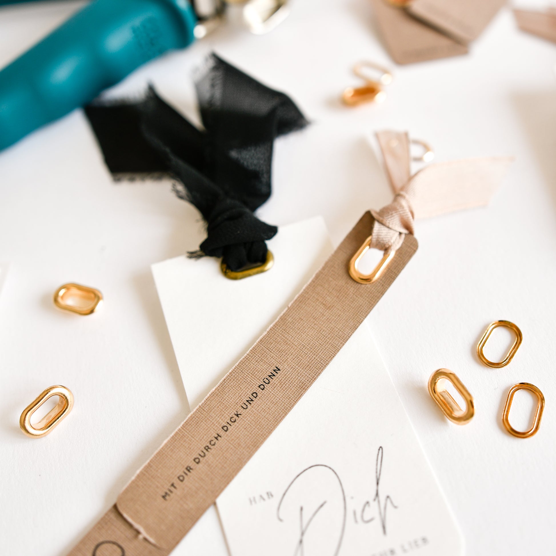 Nahaufnahme von Geschenkanhängern mit schwarzen und beigen Bändern, goldenen Ringen und Bastelzubehör – darunter der „Vaessen Crop-A-Dile“ zum Anbringen ovaler Ösen –, angeordnet auf einer weißen Unterlage.