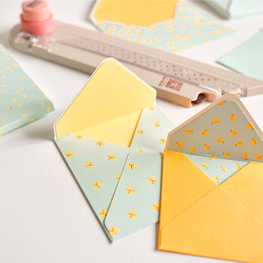 Zwei kleine, handgefertigte Umschläge aus gelbem und gemustertem Papier, die mit dem „Envelope Punch Up“ für eine präzise Falzführung hergestellt wurden, dazu Bastelwerkzeug und weiteres Papier in der Nähe – perfekt für alle, die gerne Umschläge basteln.