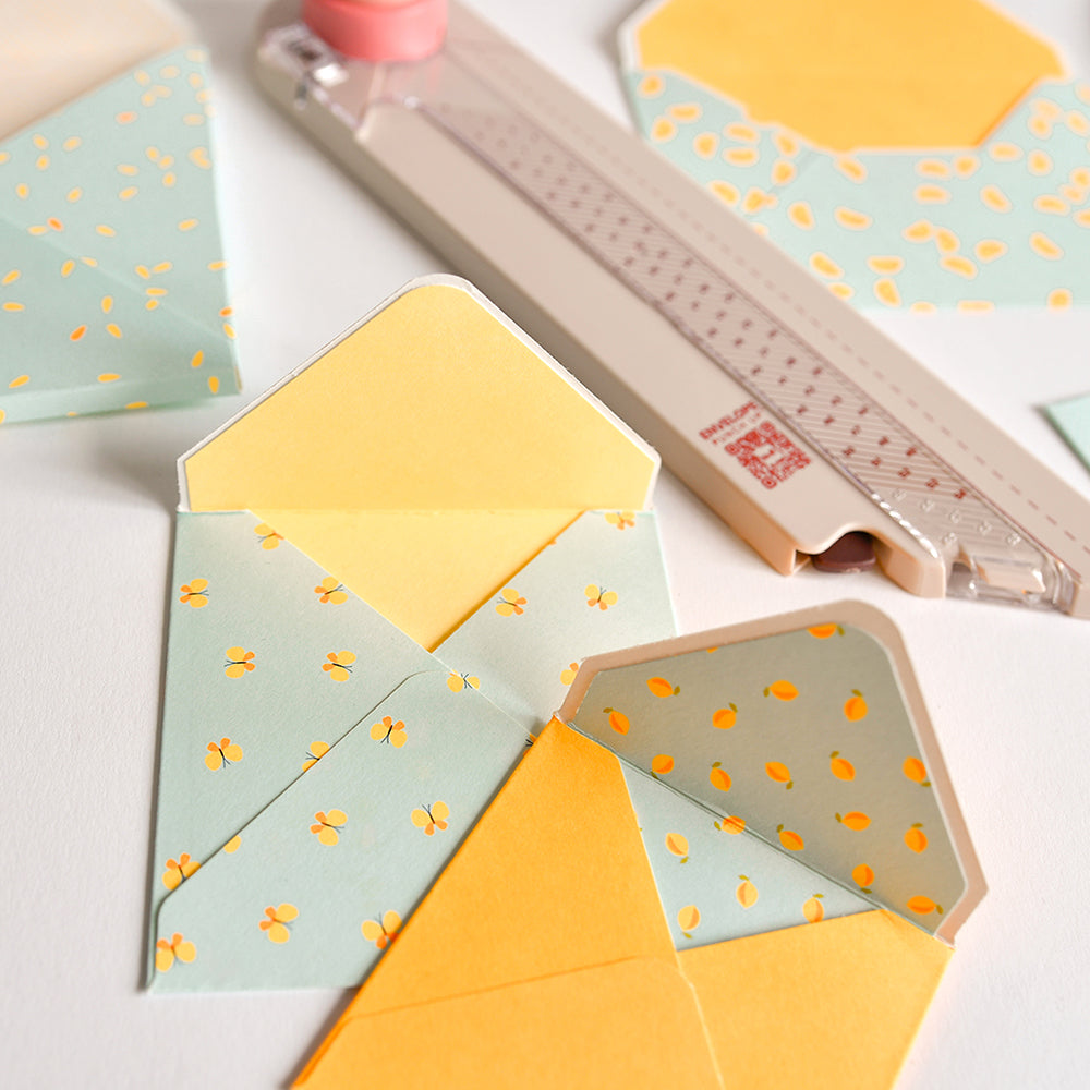 Bunte, gemusterte Umschläge und ein Papierschneider liegen auf einer weißen Unterlage bereit – perfekt für alle, die mit Hilfe eines „Envelope Punch Up“-Werkzeugs selbst Umschläge basteln möchten.
