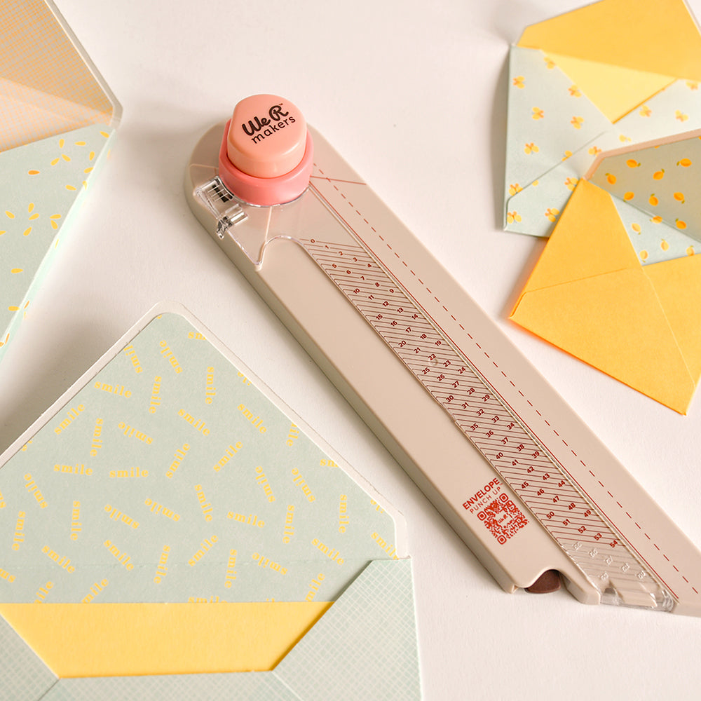 „Envelope Punch Up“-Werkzeug mit einem rosa Drehknopf, umgeben von pastellfarbenen Umschlägen mit gelb gemusterten Einlagen – ideal zum Erstellen von Umschlägen und für eine präzise Falzführung.