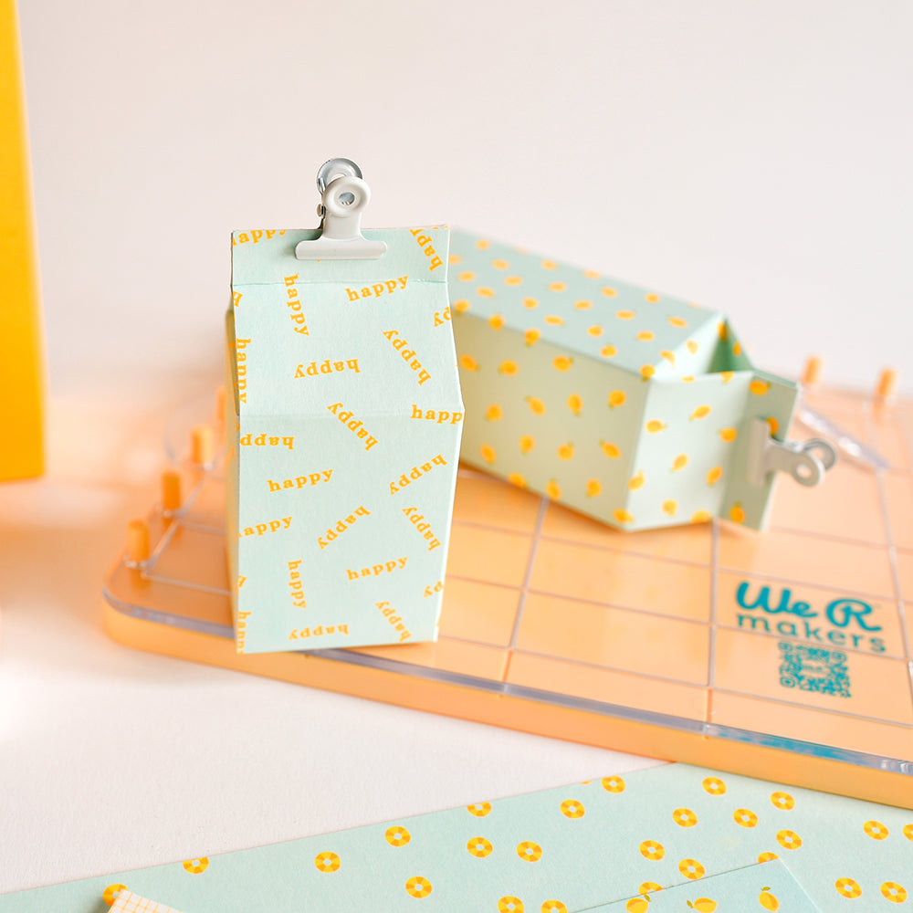 Blaue und gelbe Papierschachtel mit fröhlichem Schriftzug, mit Clipverschluss, neben einer gepunkteten Schachtel auf einer Bastelunterlage – perfekt für Papierbastelprojekte mit Ihrem Carton Stamp Board oder zum Experimentieren mit dem innovativen 3-in-1-System.