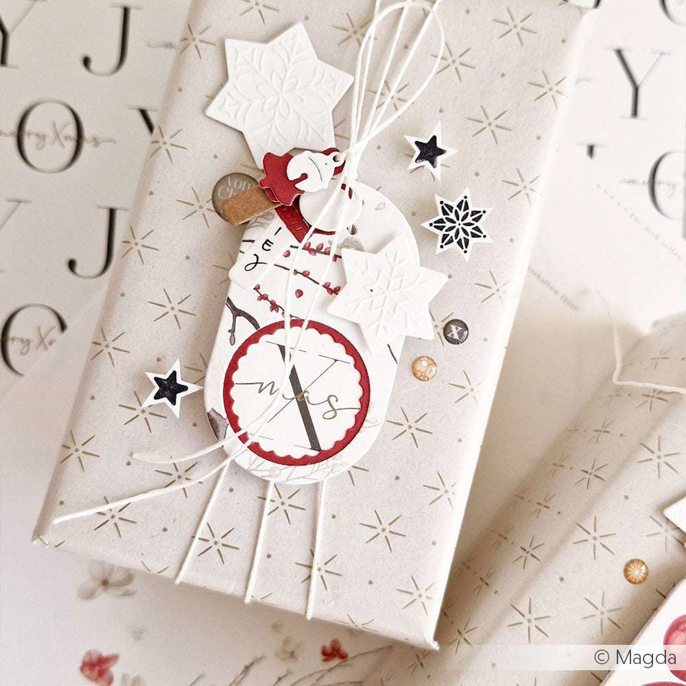 Dieses Geschenk aus weißem Sternenpapier mit festlichen Anhängern und weißer Schnur von Alexandra Renke erstrahlt vor einem weihnachtlichen Hintergrund.