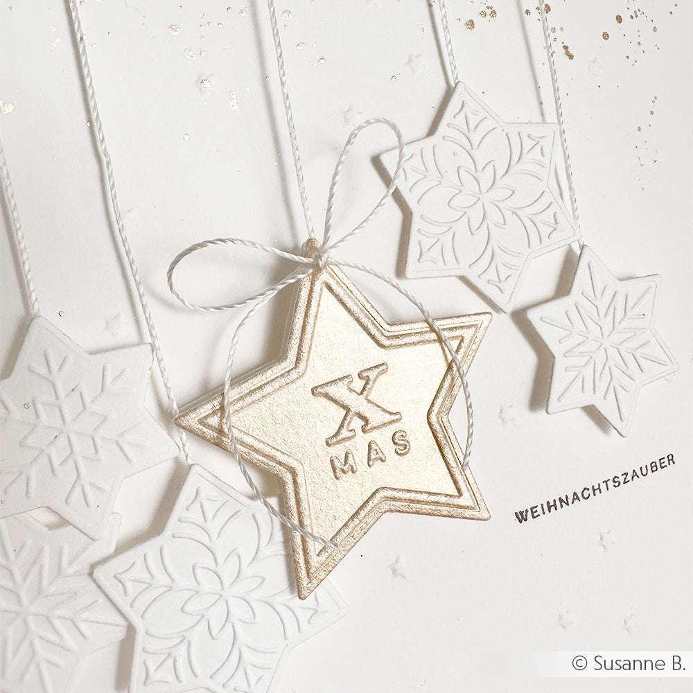 Die weiß-goldenen Sterne mit Schneeflocken und dem Text XMAS von Alexandra Renke hängen an Schnüren vor einem hellen Hintergrund - ideal, um Winterzauber in Ihre Einrichtung zu bringen.