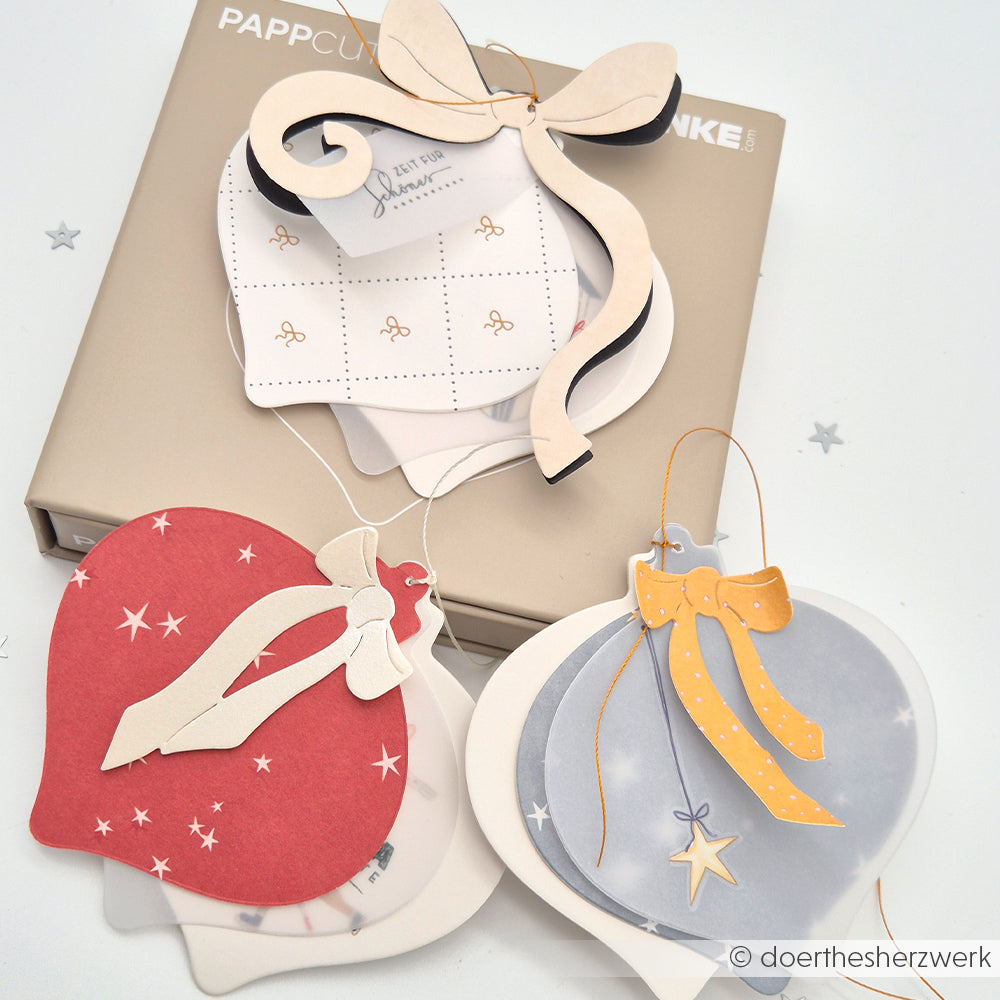 Weihnachtliche Geschenkanhänger in Form von Ornamenten in Rot, Weiß und Blau mit Schleifen, arrangiert auf einer beigen Schachtel - gebastelt von Alexandra Renke für Buchbinden oder kreative Projekte.