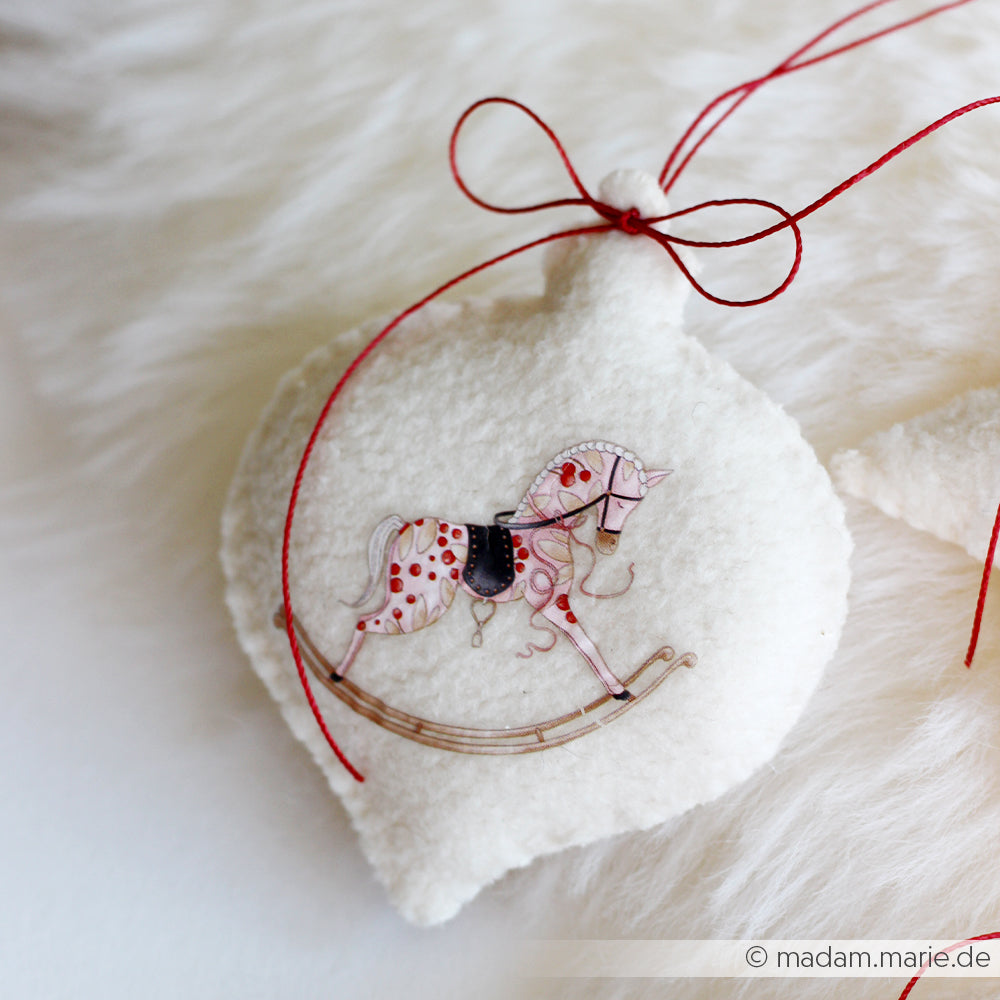 Die PAPPcut Ornamentkugel aus weißem Filz von Alexandra Renke ist mit einem gemalten Schaukelpferd und einer roten Schleife auf einer flauschigen Oberfläche verziert.