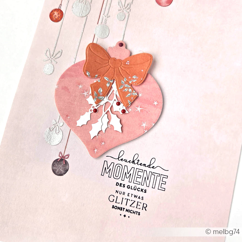 Die rosafarbene Weihnachtskarte von Alexandra Renke zeigt eine große PAPPcut Ornamentkugel mit Schleife, Stechpalme, festlichem deutschen Text und detaillierten Stahlbandtänzen als hängende Dekoration für einen eleganten Weihnachtslook.