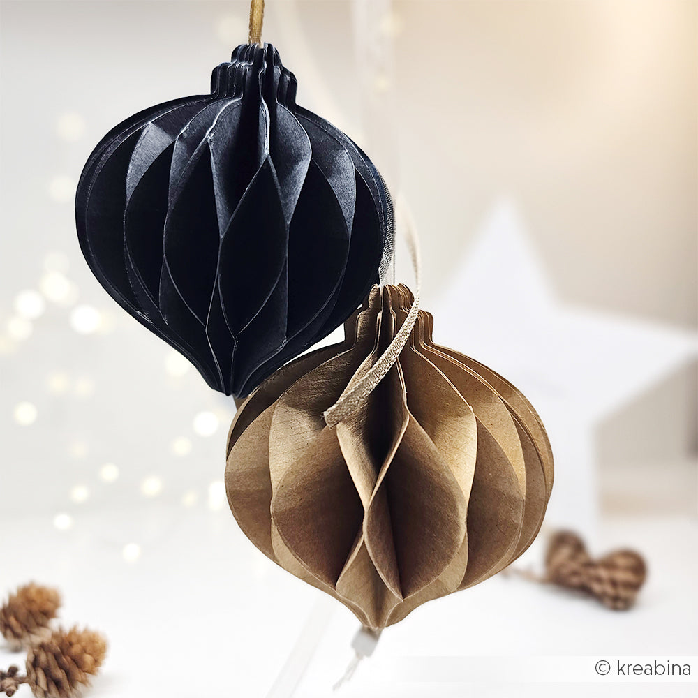 Zwei wabenförmige Papierornamente von Alexandra Renke, ein schwarzes und ein braunes, hängen anmutig mit weichen Lichtern und Tannenzapfen im Hintergrund.