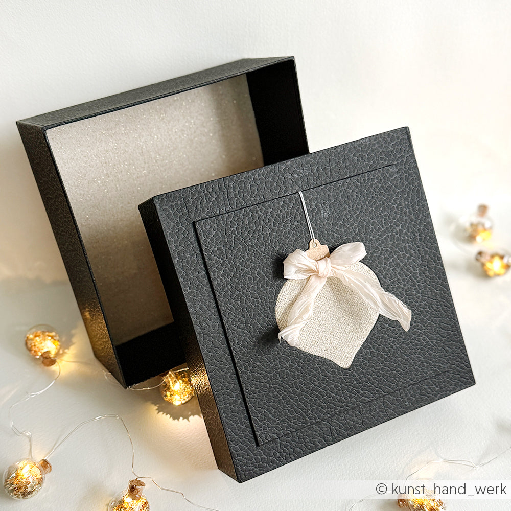 Schwarze Geschenkbox von Alexandra Renke mit strukturiertem Deckel und Schleifenverzierung, umgeben von Lichterketten - ein charmanter Akzent für Buchbinden oder kreative PAPPcuts-Projekte.