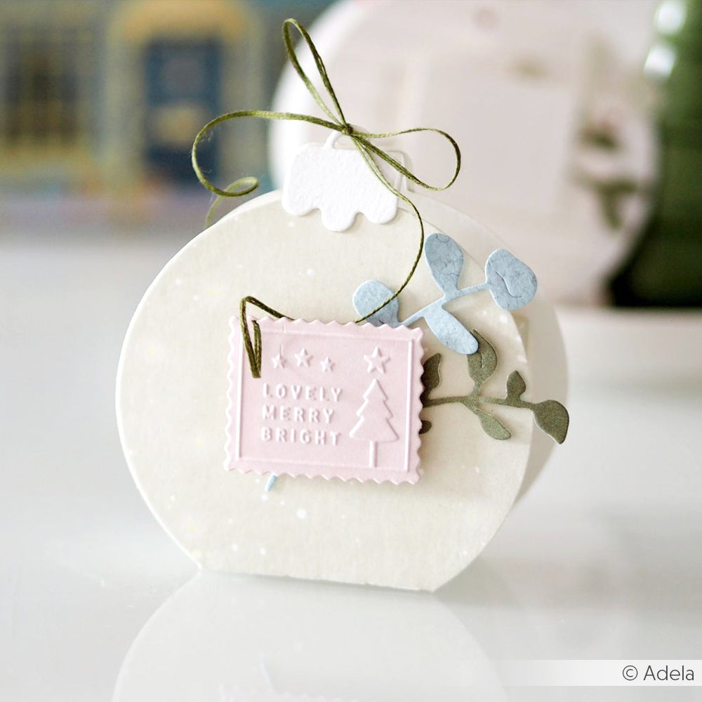 Weiße, runde Weihnachtskarte von Alexandra Renke mit einem rosafarbenen "Lovely Merry Bright"-Anhänger, grünem Band und Papierblättern - ideal zum Versenden von Weihnachtswünschen oder zum Kombinieren mit Ihren bevorzugten Cut & Emboss Folder Stanzsysteme-Designs.