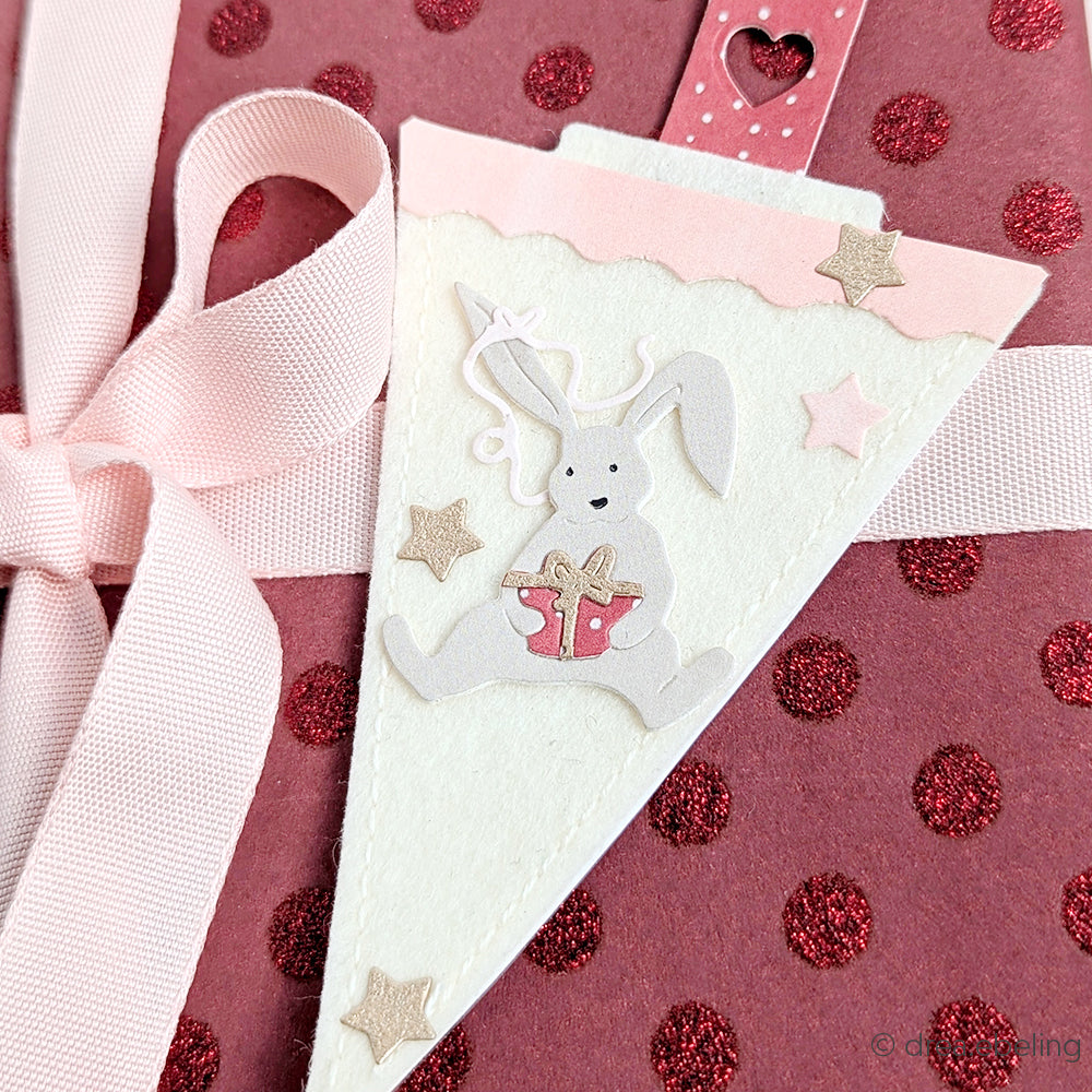 Rosa Geschenkanhänger mit einem Hasen mit Geschenk und Sternen auf einem rosa gepunkteten Hintergrund, hervorgehoben durch eine Schleife - detailgetreu mit Alexandra Renke Produkten hergestellt.