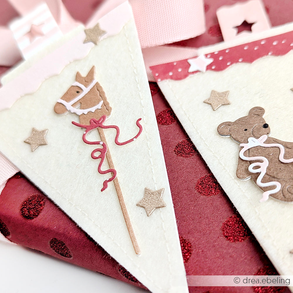 Nahaufnahme eines festlichen Papierwimpels mit einem Spielzeugpferd, einem Teddybären, Sternen und rosa Bändern - hergestellt mit Alexandra Renke-Produkten für eine besonders charmante Note.