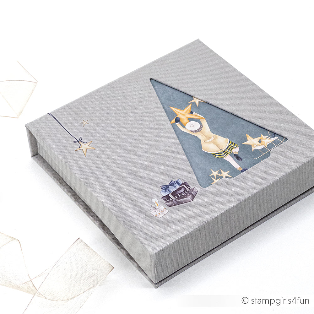 Graue Alexandra Renke-Geschenkbox mit einem ausgeschnittenen Fenster, das eine Figur zeigt, die Sterne und festliche Illustrationen platziert - ideal, um Ihre Buchbinden-Projekte oder PAPPcut Tanne-Kreationen zu präsentieren.