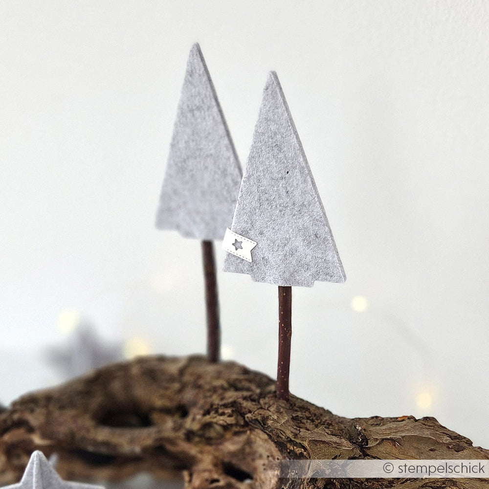 Zwei Dreiecksbäume aus grauem Filz von Alexandra Renke PAPPcut Tanne auf Stöcken sind in ein Stück Treibholz eingelassen und stehen vor einem weichen weißen Hintergrund.