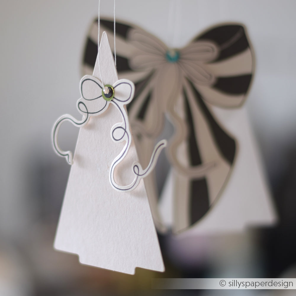 Alexandra Renkes PAPPcut Tanne Weihnachtsbaum- und Schleifenschmuck aus Papier, der mit der Buchbindentechnik hergestellt wurde, hängt vor einem verschwommenen Hintergrund.