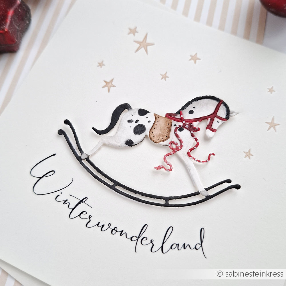 Eine Karte von Alexandra Renke mit einem gemalten Schaukelpferd, Sternen und "Winterwonderland" in Schreibschrift, alles wunderschön akzentuiert mit festlichen Rub-On Motiven.