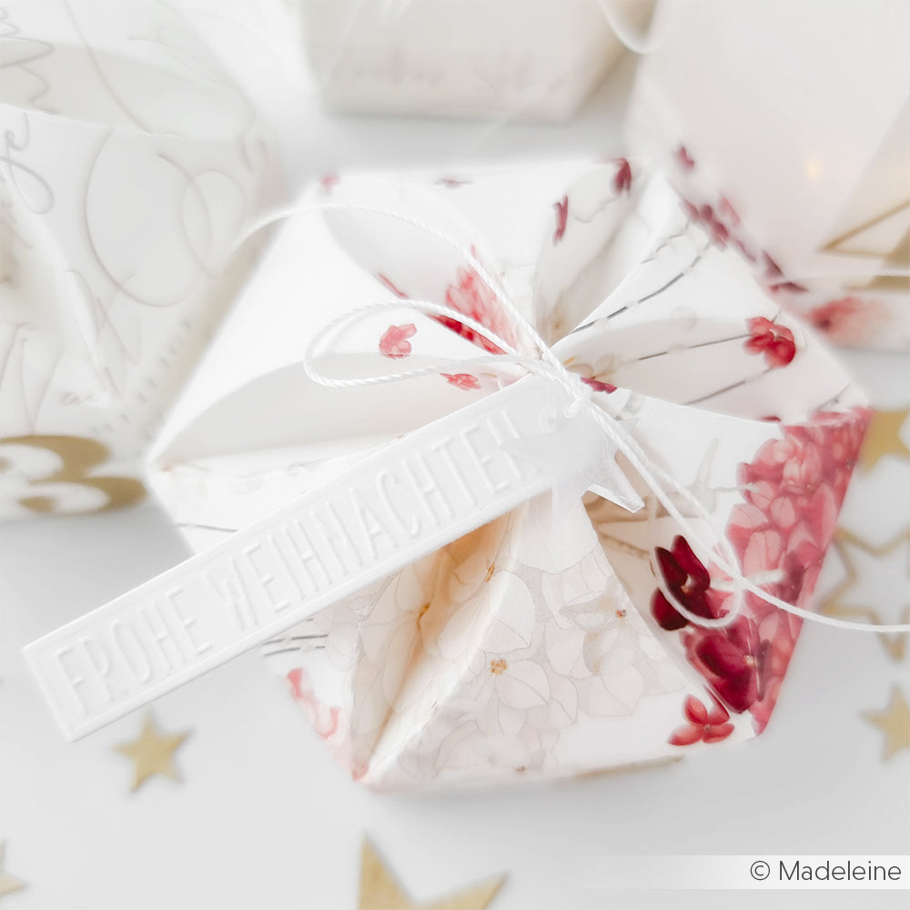 Eine floral gemusterte Geschenkbox von Alexandra Renke mit einem "Frohe Weihnachten"-Anhänger und weißer Kordel, gefertigt aus elegantem Designpapier und verziert mit Sternen.