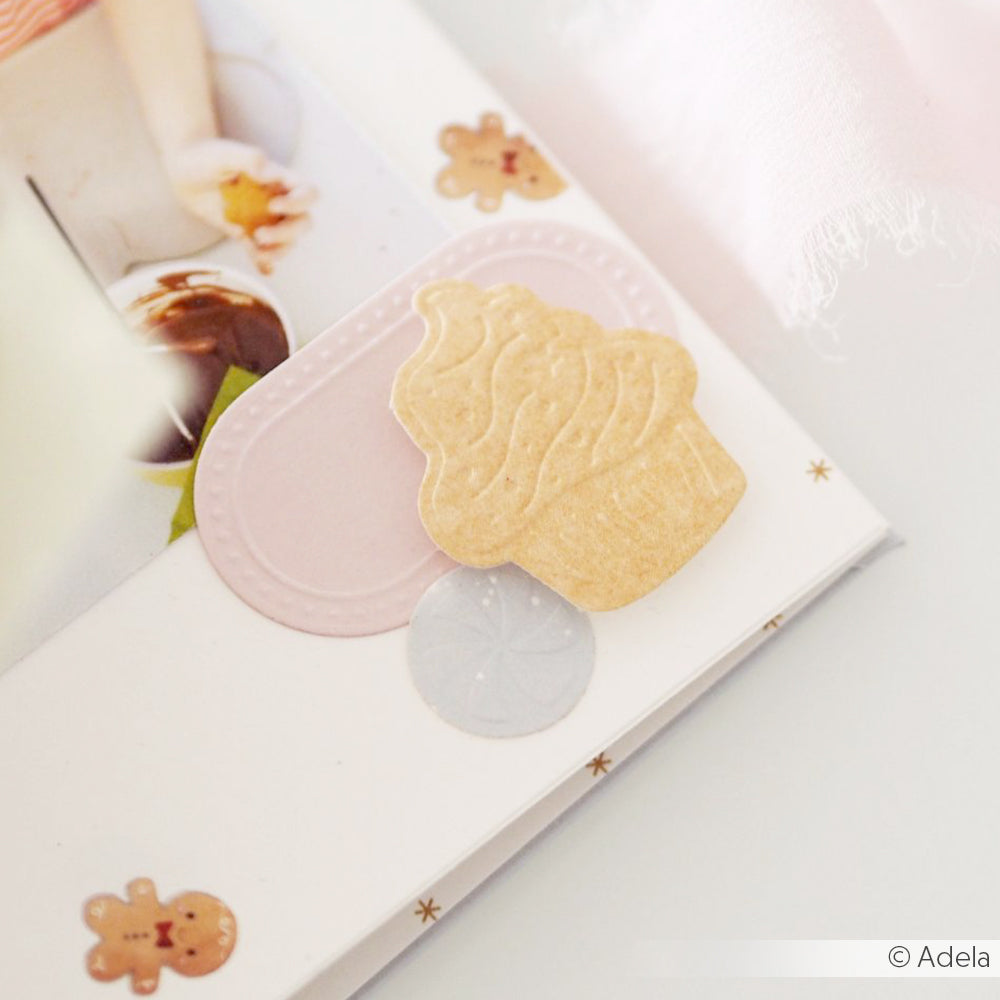Scrapbook-Seite mit Aufklebern in Cupcake- und Pastellfarben, Lebkuchenmännern und einer Kinderhand im Hintergrund, mit zarten Akzenten, die mit Alexandra Renke Cut & Emboss Folder-Designs hergestellt wurden und für eine charmante Note sorgen.