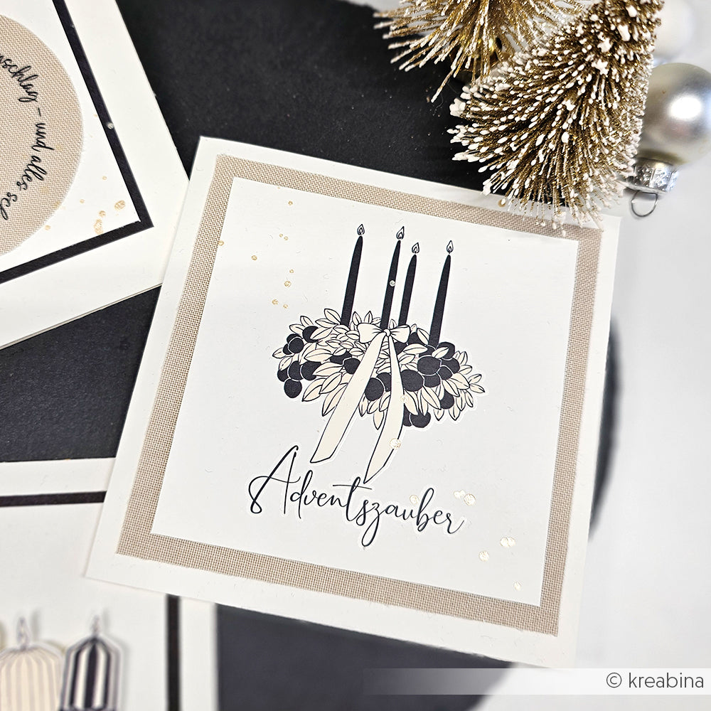 Eine Karte von Alexandra Renke mit vier schwarzen Kerzen, einem floralen Muster, dem Wort "Adventszauber" darunter und Rub-On Festlichkeiten neben goldenen Weihnachtsdekorationen.