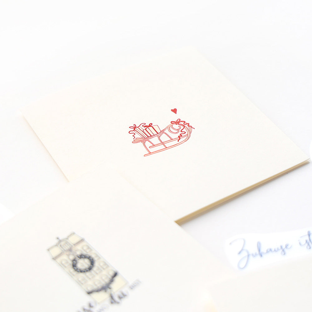 Auf dieser cremefarbenen Karte von Alexandra Renke ist ein roter Schlitten mit Geschenken und einem Herz abgebildet - ideal für Bastelprojekte oder um Ihre Weihnachtsbasteleien mit festlichen Rub-On Motiven zu versehen.