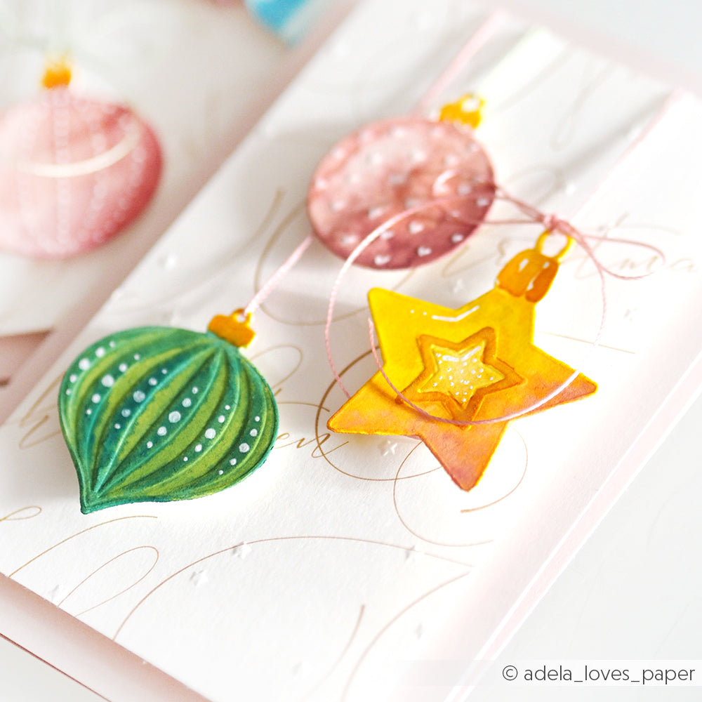 Drei farbenfrohe Festtagskugeln - grün, rosa und gelb - zieren eine weiße, gemusterte Karte, die Alexandra Renke mit Cut & Emboss Folder-Techniken gestaltet hat.