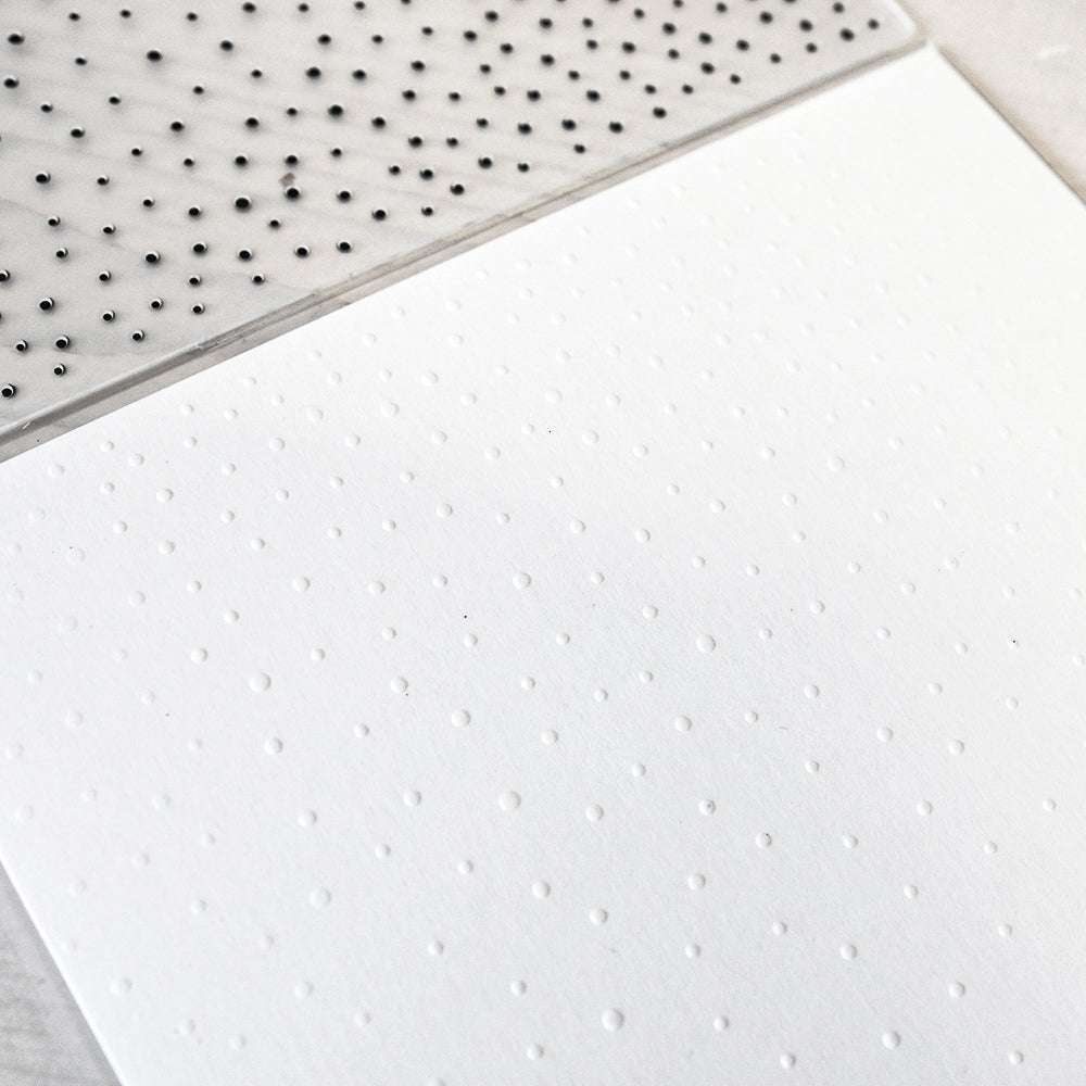 Ein weißes Blatt, das mit dem erhabenen Punktmuster Leichter Schneefall von Alexandra Renke geprägt ist, liegt neben einem klaren Prägefolder mit schwarzen Punkten.