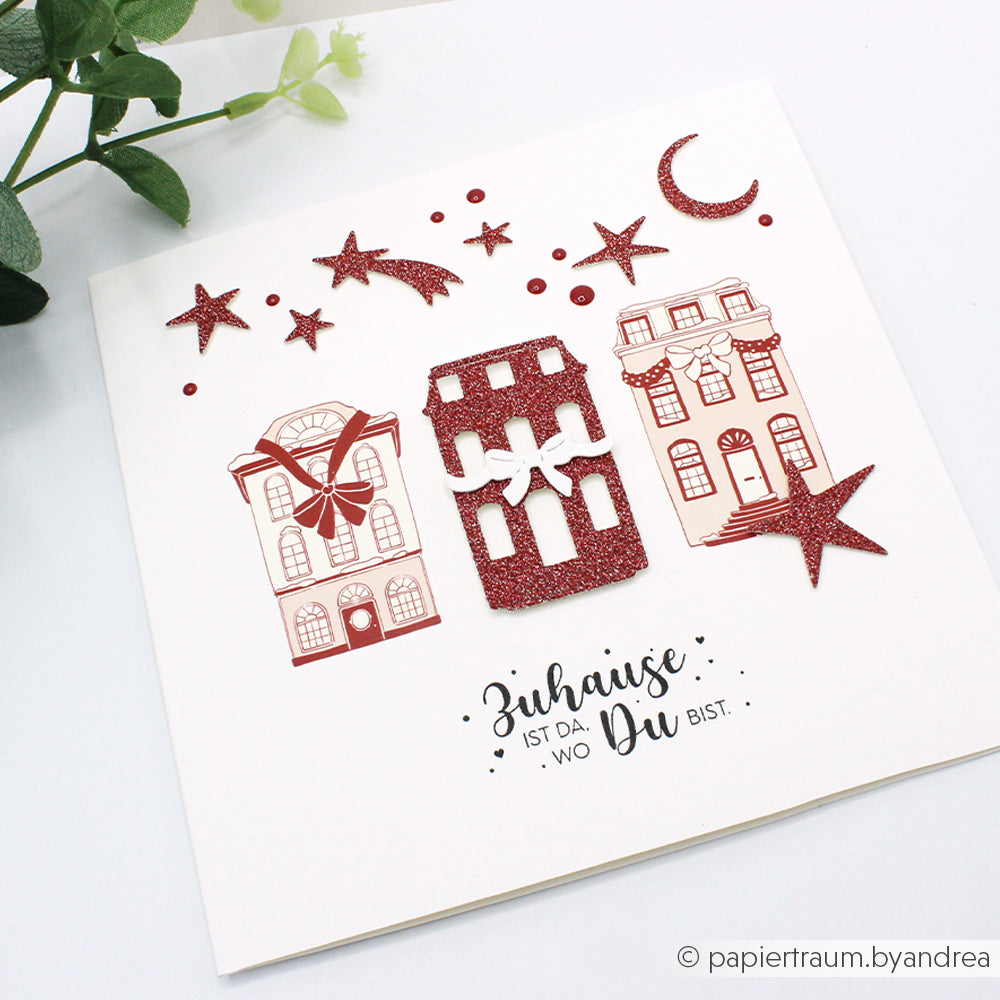Karte von Alexandra Renke mit drei roten Häusern, Glitzersternen, einem Mond und dem deutschen Text "Zuhause ist da, wo Du bist". Ideal für winterliche Bastelprojekte und festliche Dekorationen mit Transferfolien und Rubbelfolien.