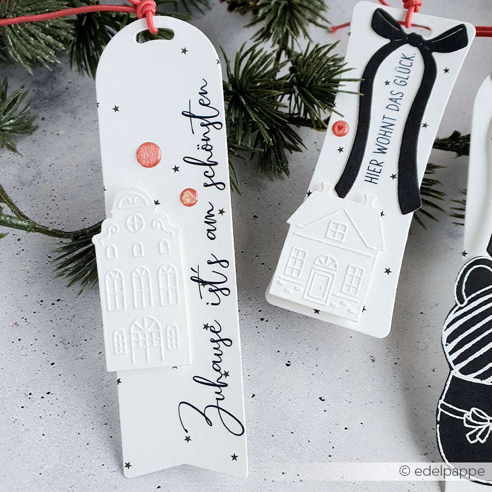Weiße Alexandra Renke Lesezeichen mit geprägten Hausmotiven und deutschem Text, die an roten Schnüren mit Tannenzweigen hängen - ideal für Motive Winterdeko oder Rub-On Festlichkeiten Winterheim Bastelprojekte.