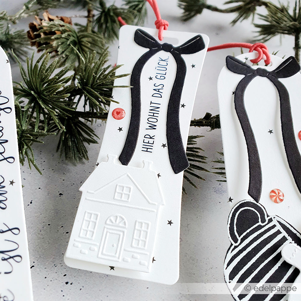 Das weiße Lesezeichen von Alexandra Renke mit schwarzer Schleife, kleinem Hausmotiv und deutschem Text - hergestellt mit Rub-On Festlichkeiten Winterheim - ziert einen Tannenzweig. Ideal für Bastelprojekte Winter oder Transferfolien Basteln.