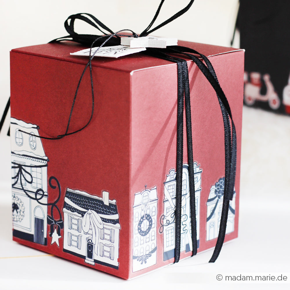 Roter Geschenkkarton mit schwarzem Band und festlichen Häusern auf dem Boden - dieses Stück von Alexandra Renke ist perfekt für Bastelprojekte Winter und kann mit Rub-On Festlichkeiten Winterheim personalisiert werden.