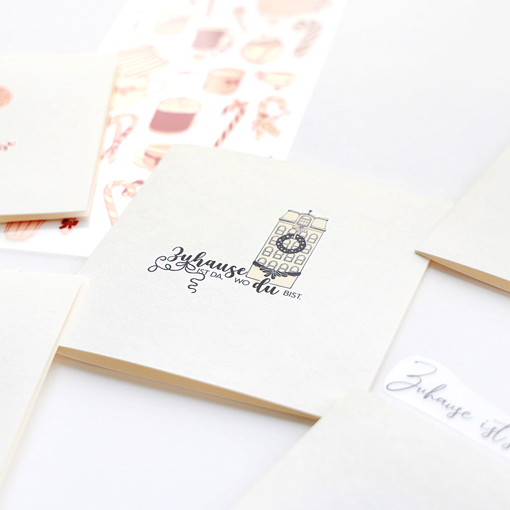 Quadratische Grußkarten von Alexandra Renke mit deutschem Text und Bau-Illustrationen, umgeben von Zusatzkarten, dekorativen Aufklebern und kreativen Motiv-Transfer-Designs für Rub-On Festlichkeiten Winterheim.