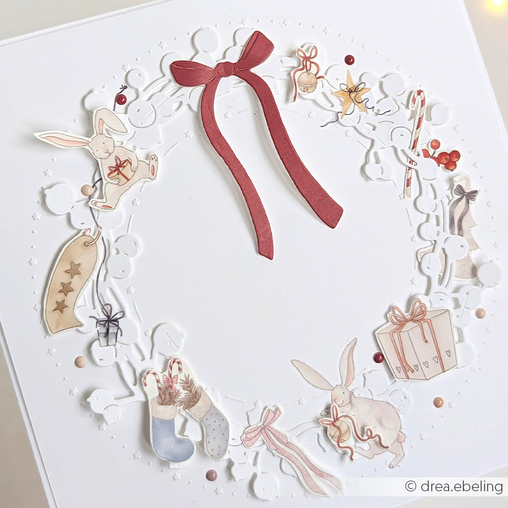 Weiße Karte von Alexandra Renke mit dekorativem Kranz, roter Schleife, Häschen, Strümpfen und Geschenken - ideal zum Aufstellen von Figurenwelt-Motiven oder zum Personalisieren mit Rub-On Transferfolien.