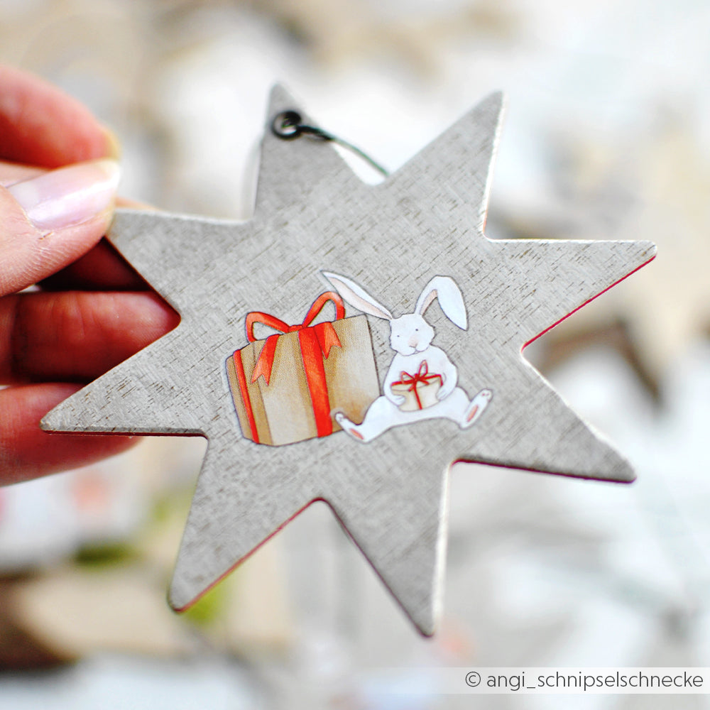 Eine Hand hält ein sternförmiges Ornament mit einem Hasen und einem Geschenkdesign von Alexandra Renke - ideal für Bastelprojekte Weihnachten oder zum Verzieren mit Rub-On Weihnachtszauber-Techniken.