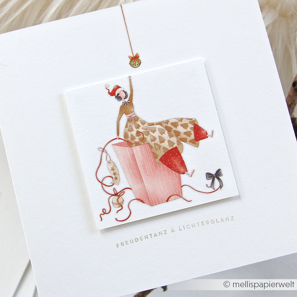 Illustration einer fröhlichen Frau, die auf einer rosafarbenen Geschenkschachtel mit festlichen Bändern und Schleifen sitzt, inspiriert von Alexandra Renke Transferfolien Basteln und Rub-On Motiven für zusätzlichen handwerklichen Weihnachtscharme.