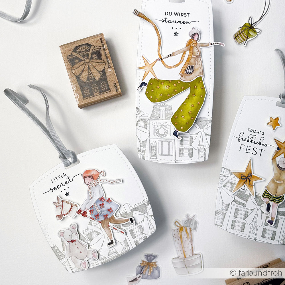 Alexandra Renke präsentiert drei festliche Geschenkanhänger mit skurrilen Weihnachtsillustrationen, deutschen Grüßen und Schleifen - ideal für Transferfolien-Bastelprojekte oder um Weihnachtsmotive mit Rub-On-Charme in Ihre Weihnachtsgeschenke zu integrieren.