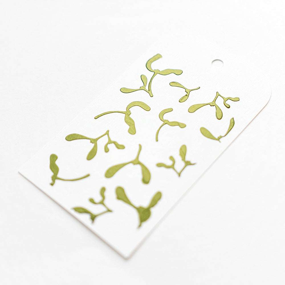 Ein weißer Anhänger mit grünen botanischen Zweigen, erstellt mit wiederverwendbaren Schablonen von Alexandra Renke, dargestellt auf einem klaren weißen Hintergrund.