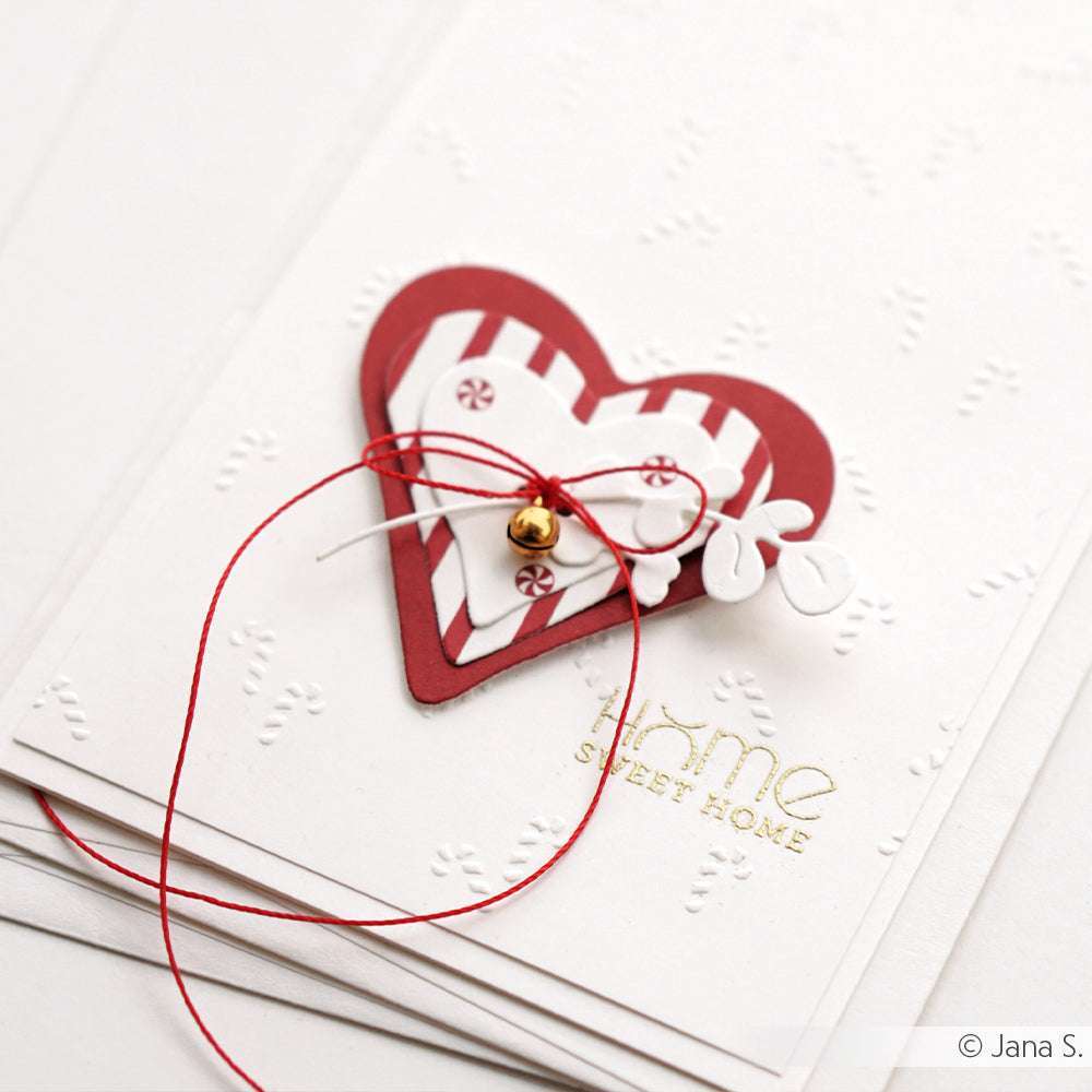 Handgefertigte Karte von Alexandra Renke mit einem rot gestreiften Herz, inspiriert von Zuckerstangen-Gewusel, weißen Blättern, roter Schnur und goldenem Text "Home sweet home". Der Embossing-Folder verleiht der Karte ein charmantes Finish mit strukturierten Details.