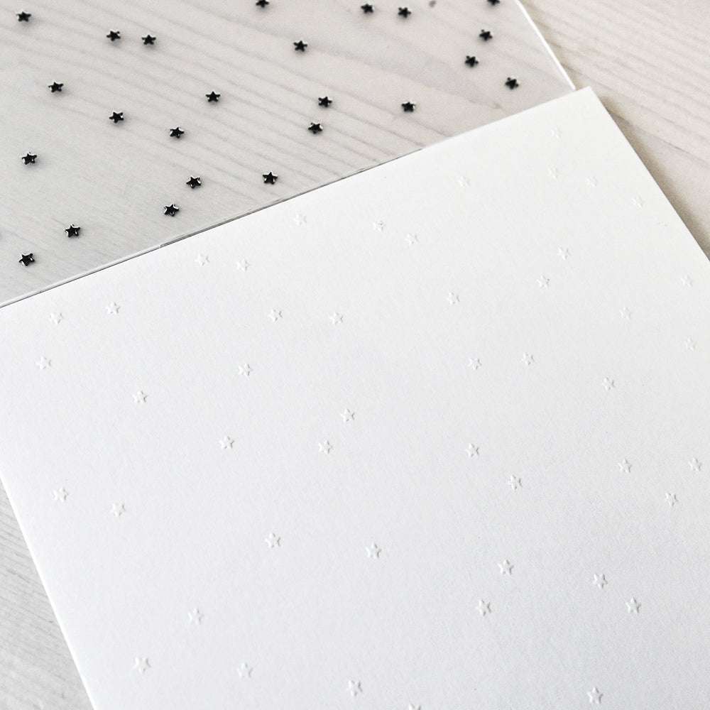 Transparente Folie mit schwarzem Sternenmuster auf weißem Alexandra Renke Prägen Papier, mit dezent geprägten Sternen, die mit einem Embossing Folder erstellt wurden, auf einer hellen Oberfläche.