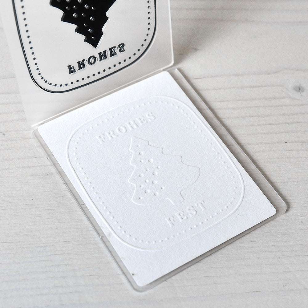 Weiße Karte von Alexandra Renke mit einem Weihnachtsbaum, der mit einem Prägefolder hergestellt wurde, und dem deutschen Gruß "Frohes Fest", der auf einer hellen Holzoberfläche dargestellt ist.