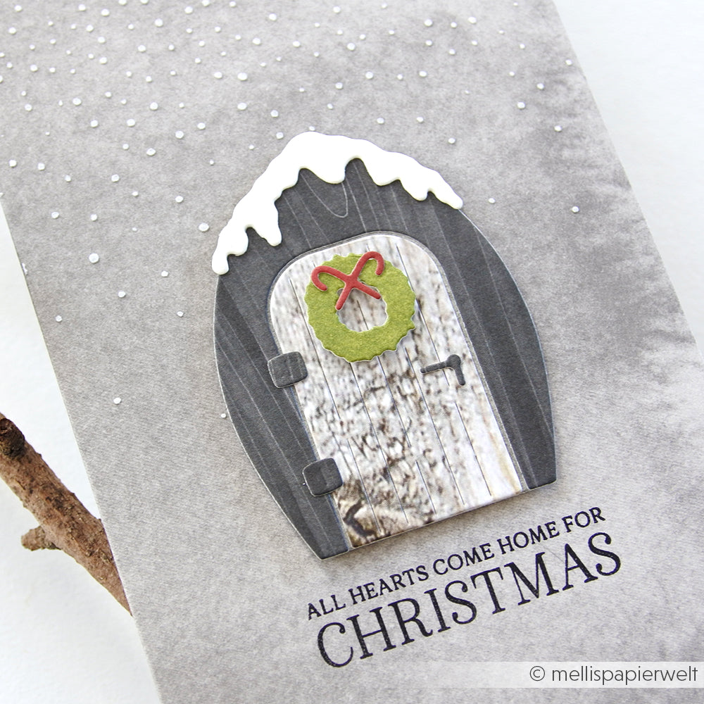Eine verschneite Tür mit einem grünen Kranz ziert diese graue Karte von Alexandra Renke, die mit der wiederverwendbaren Schablone Schneefall gestaltet wurde. Der Text lautet: Alle Herzen kommen zu Weihnachten nach Hause.