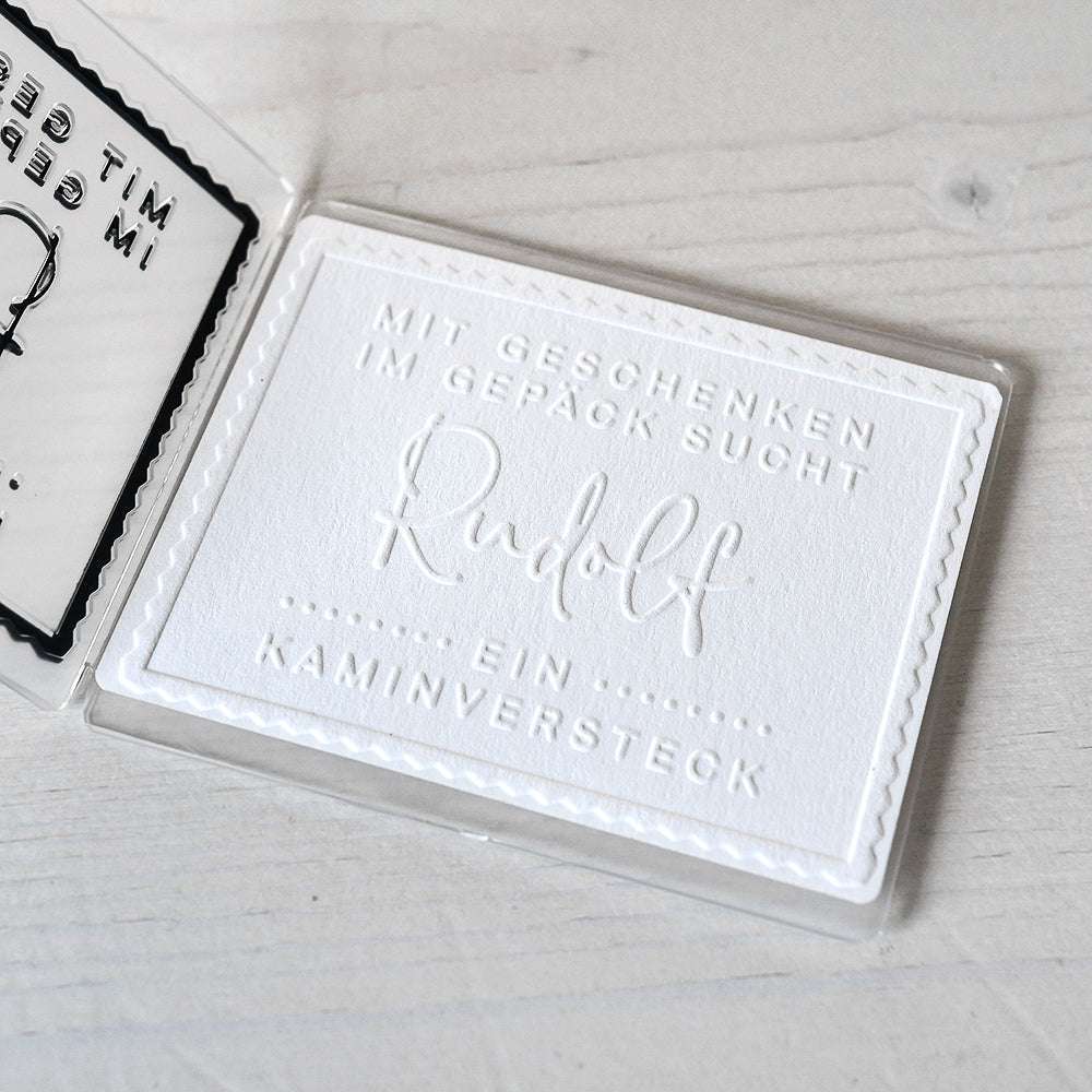 Weiße Prägekarte von Alexandra Renke mit deutschem Text: "Mit Geschenken im Gepäck sucht Rudolf ein Kaminversteck." Hergestellt mit Designpapier und Prägefolder für eine elegante, festliche Note für Rudolfs Geschenke.