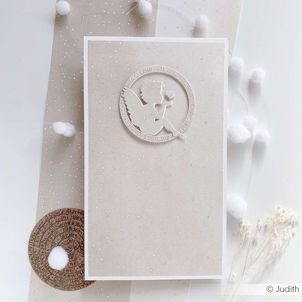 Minimalistische Karte von Alexandra Renke mit einer weißen Engelssilhouette in einem Kreis auf beigefarbenem, schneebedecktem Papier, akzentuiert durch weiße Bommeln. Ideal für die Verwendung mit Stahlstempeln oder Stanzsystemen.