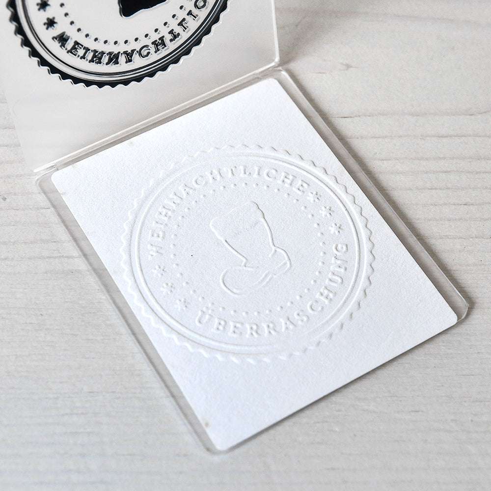 Weiße, mit dem Alexandra Renke Prägefolder geprägte Karte mit dem kreisförmigen Stempel eines Weihnachtsstrumpfs und dem deutschen Text "Weihnachtliche Überraschung". Ideal für festliche Grüße auf Ihren Lieblingsdesignpapieren.