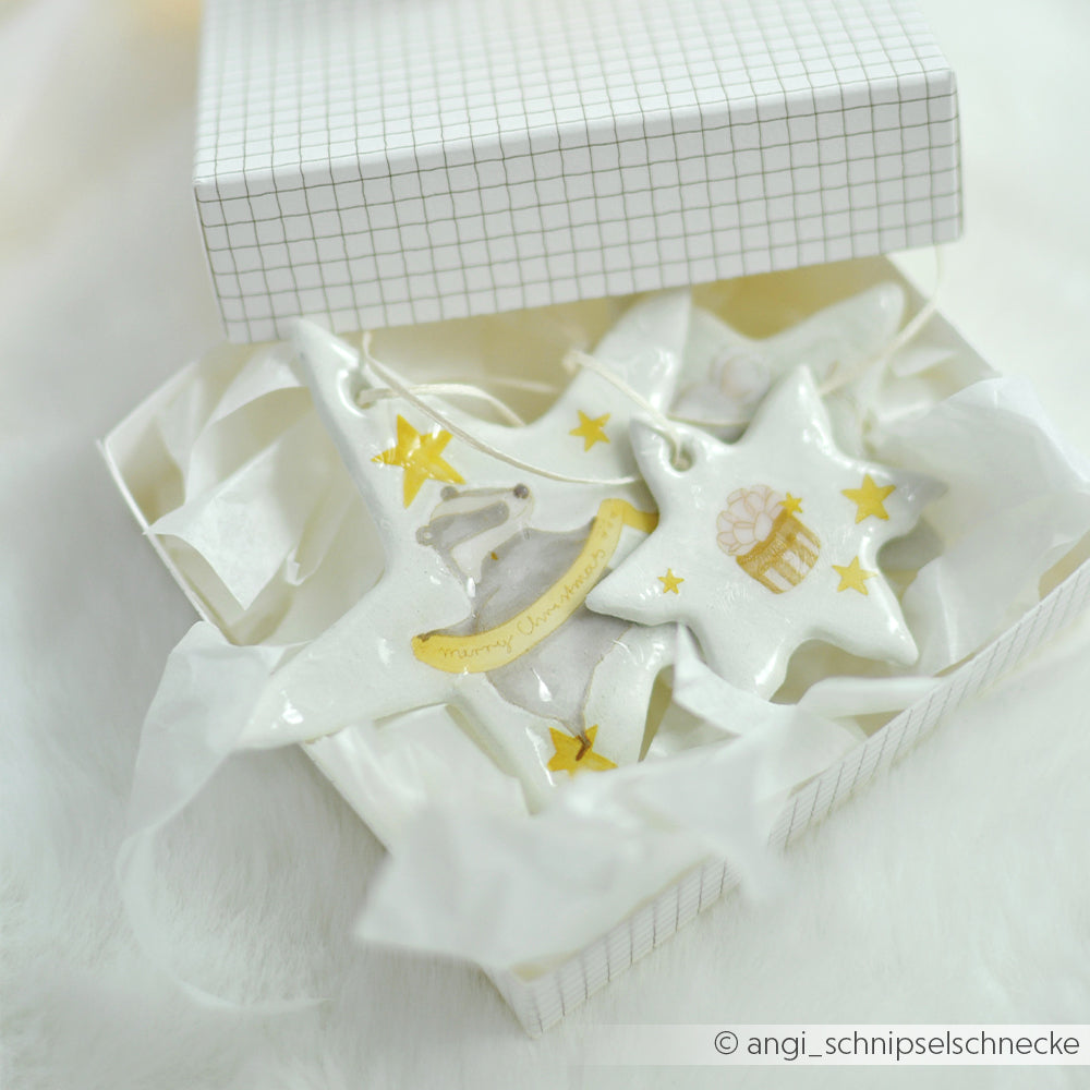 Zwei sternförmige Keramikornamente, verziert mit Rub-On Weihnachtswald, verpackt in einer Geschenkbox.