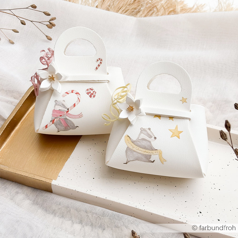 Zwei Geschenktüten aus weißem Papier mit Rub-On-Motiven aus der Weihnachtswald-Reihe: Dachs-Illustrationen, Blumen und Bänder.