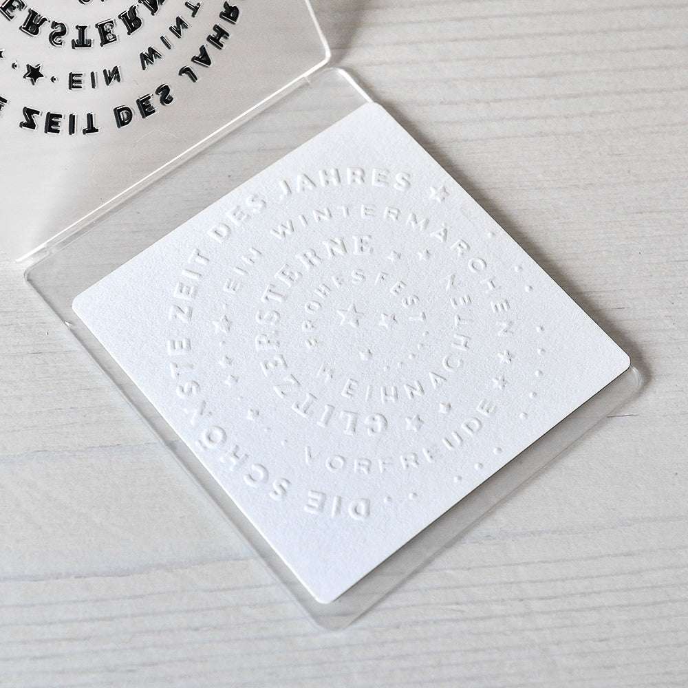 Weiße Karte von Alexandra Renke, die mit einem Prägefolder hergestellt wurde und den geprägten deutschen Text "Typo Kreis Schönste Zeit" in konzentrischen Kreisen auf einer hellen Holzoberfläche zeigt.