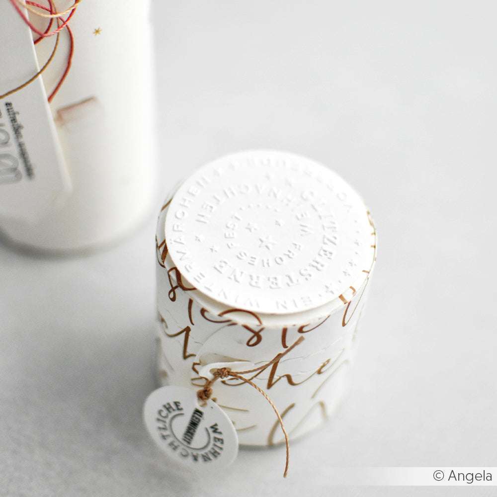 Die weiße, zylindrische Schachtel von Alexandra Renke mit goldener Schrift und einem runden, geprägten Deckel, der an einen Embossing Folder erinnert, ist aus Designpapieren gefertigt und ruht auf einer hellen Oberfläche.