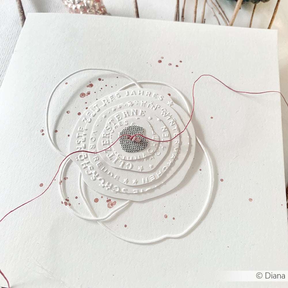 Weiße, mit dem Alexandra Renke Prägefolder geprägte Karte mit einem Herzanhänger, rosa Faden und rosa Farbspritzern auf weißem Hintergrund.