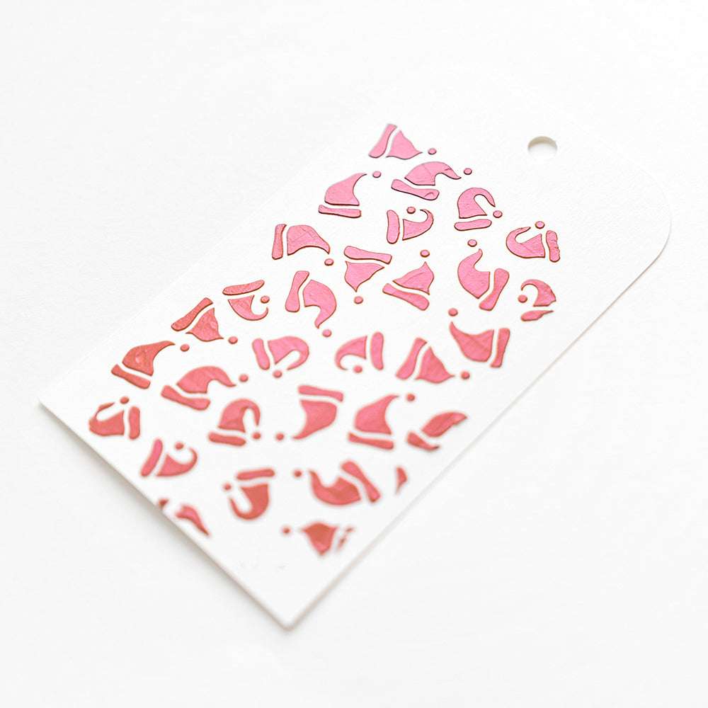 Weißer Geschenkanhänger von Alexandra Renke, verziert mit rosafarbenen Nikolausmützen und einem Loch oben für die Schnur, hergestellt mit Stencil Nikolausmützen Schablonen. Perfektes wiederverwendbares Accessoire für festliche Geschenke.